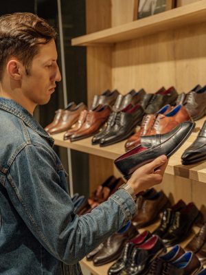 A man in denim jacket selects leather shoes from a store shelf indoors.